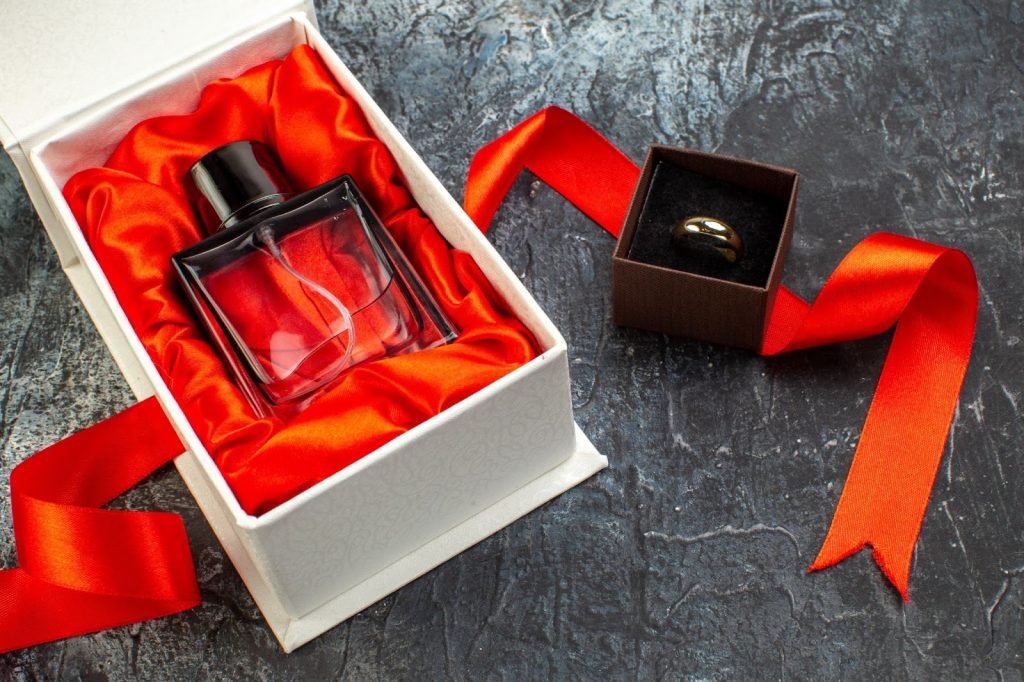 Open a gift box with red satin, holding a cologne bottle, beside a ring box, and flowing red ribbon on a dark stone surface.
